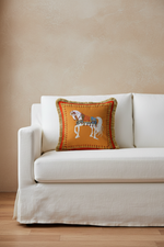 A styled armchair in a bright room with sheer white curtains, featuring a decorative mustard yellow embroidered throw pillow with a horse design. A woven side table with pink roses and a floor lamp are positioned next to the chair.