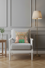 A light beige slipcovered loveseat sofa with a decorative throw pillow featuring a colorful architectural arch design, set against a textured beige wall and light wood floor.