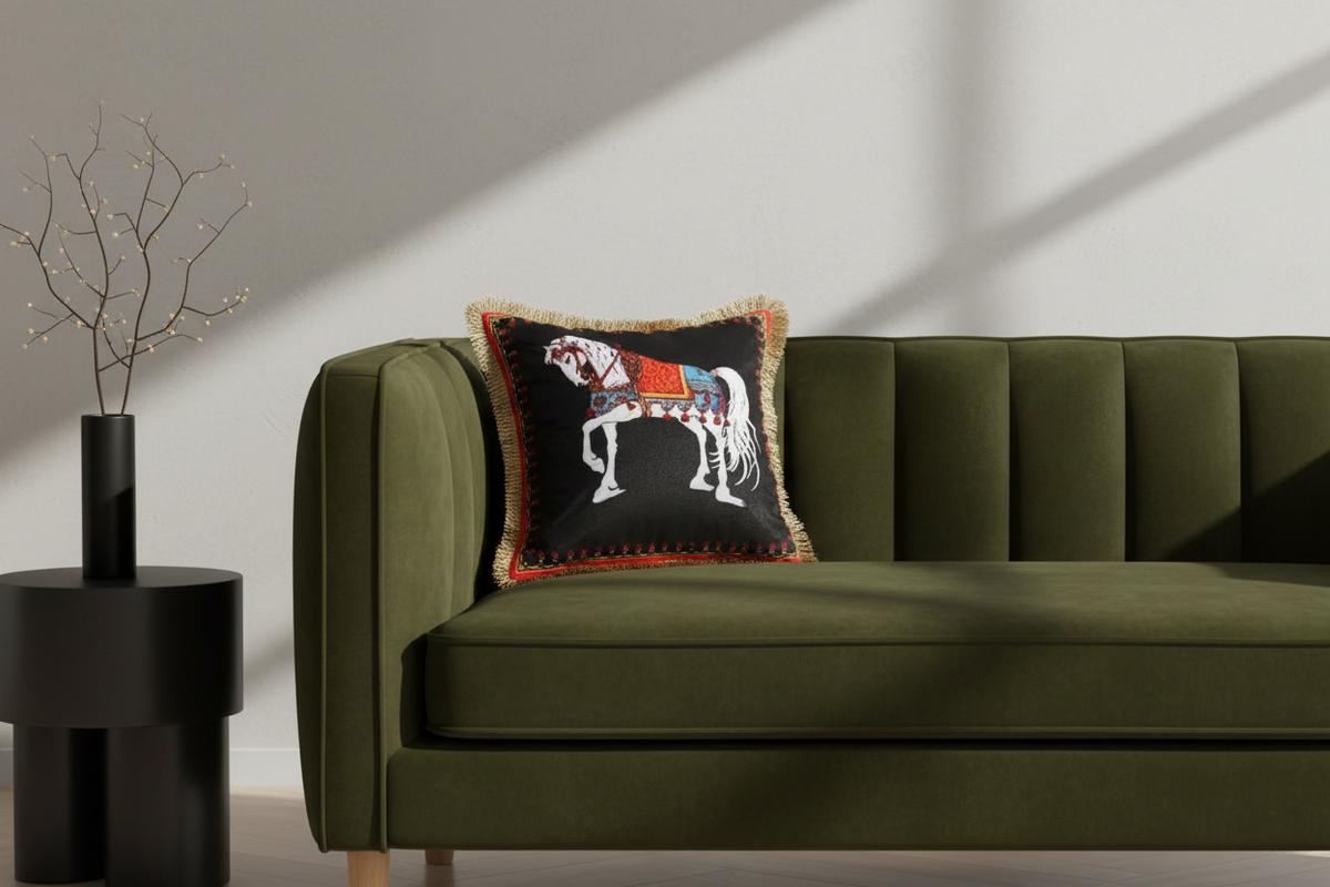 A square black throw pillow with a white embroidered horse and colorful saddle details, styled on a white slipcovered sofa against a beige wall.