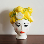 Decorative vase shaped like a woman's head with a yellow floral arrangement on a wooden surface.