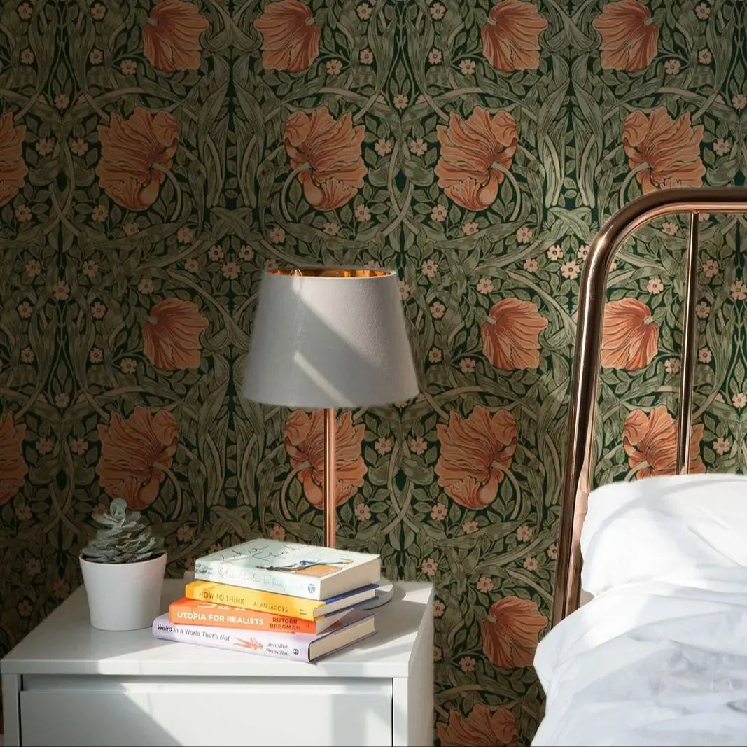 Room interior with floral wallpaper, lamp, books, and a plant on a nightstand.