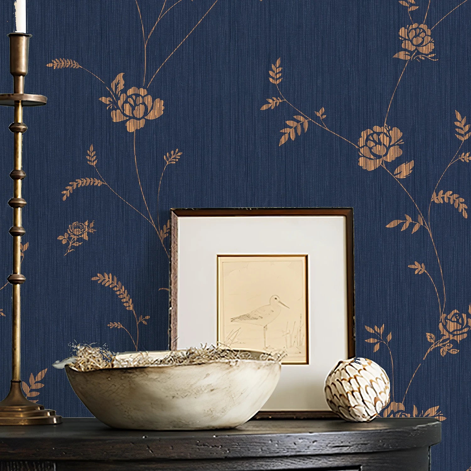 Decorative setting with a bowl, candlestick, and framed picture against a floral-patterned blue wall.