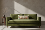 An olive green velvet loveseat sofa with a floral accent pillow, set against a textured gray wall with dramatic diagonal sunlight shadows. A small wooden side table with a vase of dried branches stands to the left.