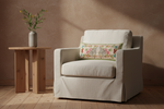 A cream-colored slipcovered armchair with a decorative floral throw pillow, set in a rustic room next to a small wooden side table with a vase of greenery.