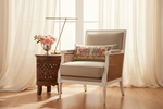 A classic-style armchair with a beige upholstered seat and back, a white frame, and woven cane armrests, styled with a floral accent pillow. It is set in a bright room with sheer cream curtains, a wooden floor, and a matching carved wooden side table holding a vase of pink roses and a gold floor lamp.