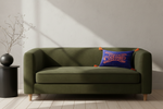An olive green fabric loveseat sofa with light wood legs, styled with a blue decorative throw pillow in a minimalist room with a black side table and dried branches.