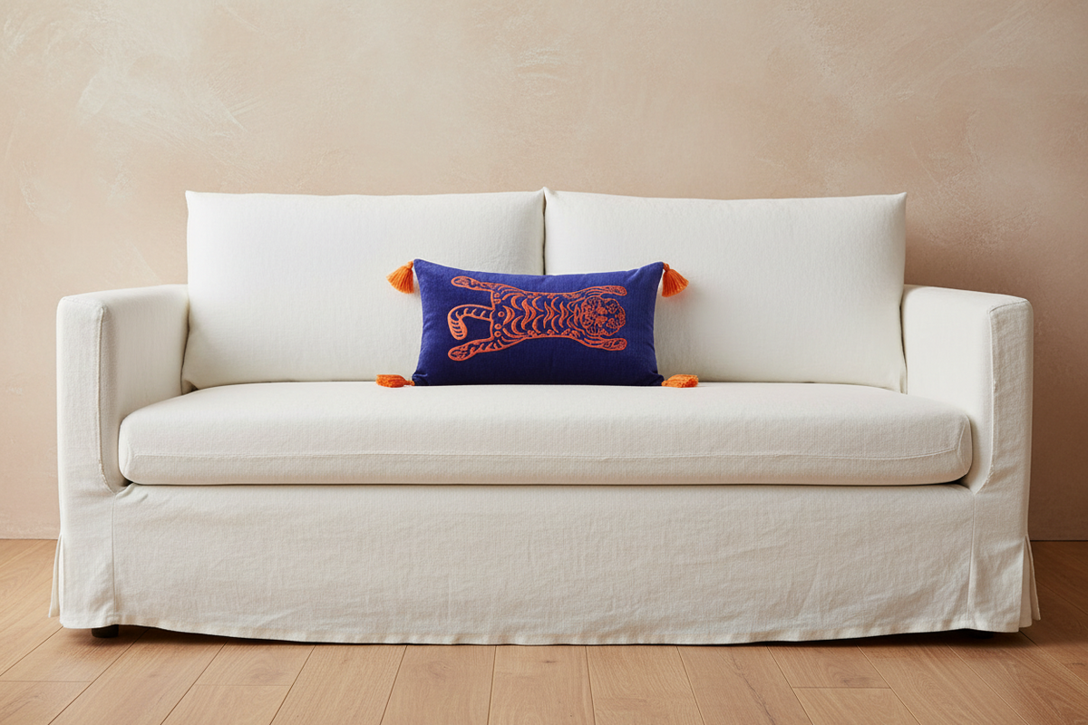 A white slipcovered sofa with a deep skirt sits against a warm beige textured wall on light wood flooring. A small blue throw pillow with orange embroidered details and tassels rests on the sofa.