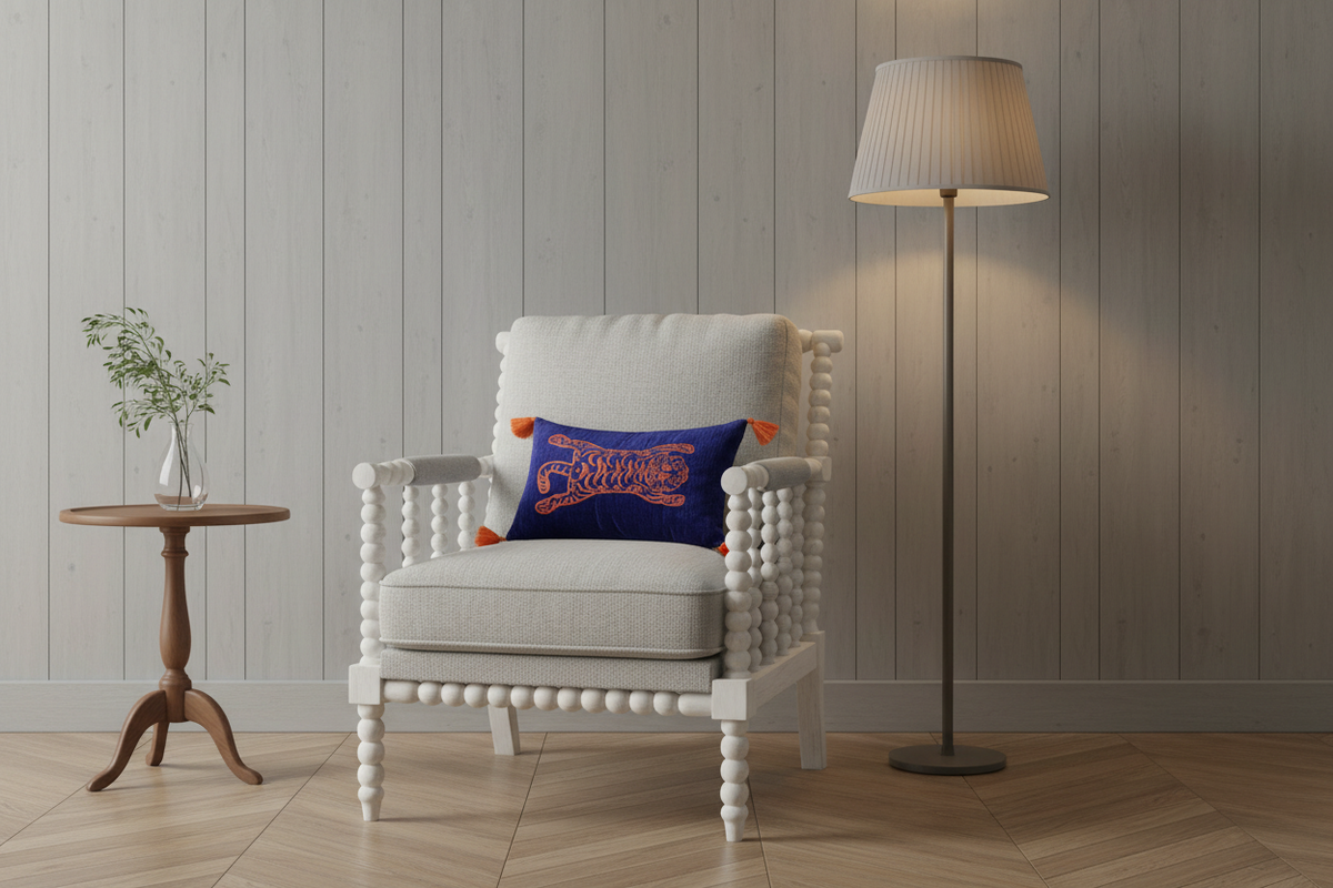 A white wooden armchair with bead-style turned legs and a white cushion, styled in a room with a gray wood-paneled wall, a floor lamp, a small wooden side table with a vase of greenery, and a blue throw pillow featuring orange text.