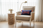 A white and woven rattan armchair with cream cushions sits in a bright room with sheer white curtains, a carved wooden side table with flowers, a gold floor lamp, and a blue throw pillow with orange embroidery.