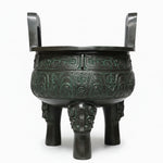 Bronze tripod cauldron with intricate designs on a wooden stand against a gray background.
