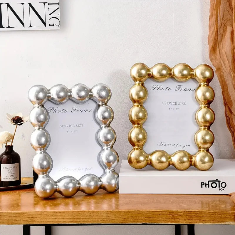 Two decorative photo frames, one silver and one gold, on a wooden surface with a white wall in the background.