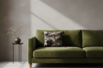 An olive green velvet sofa in a modern living room setting, styled with a dark patterned accent pillow and a small side table holding a vase with dried branches against a textured gray wall.