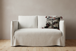 A cream-colored slipcovered loveseat sofa with a textured linen-like fabric, shown with a patterned accent pillow on a light wood floor against a neutral beige wall.