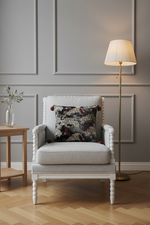 A light gray upholstered armchair with a dark patterned accent pillow, set in a styled room with a gray paneled wall, a wooden side table, a floor lamp, and a herringbone wood floor.
