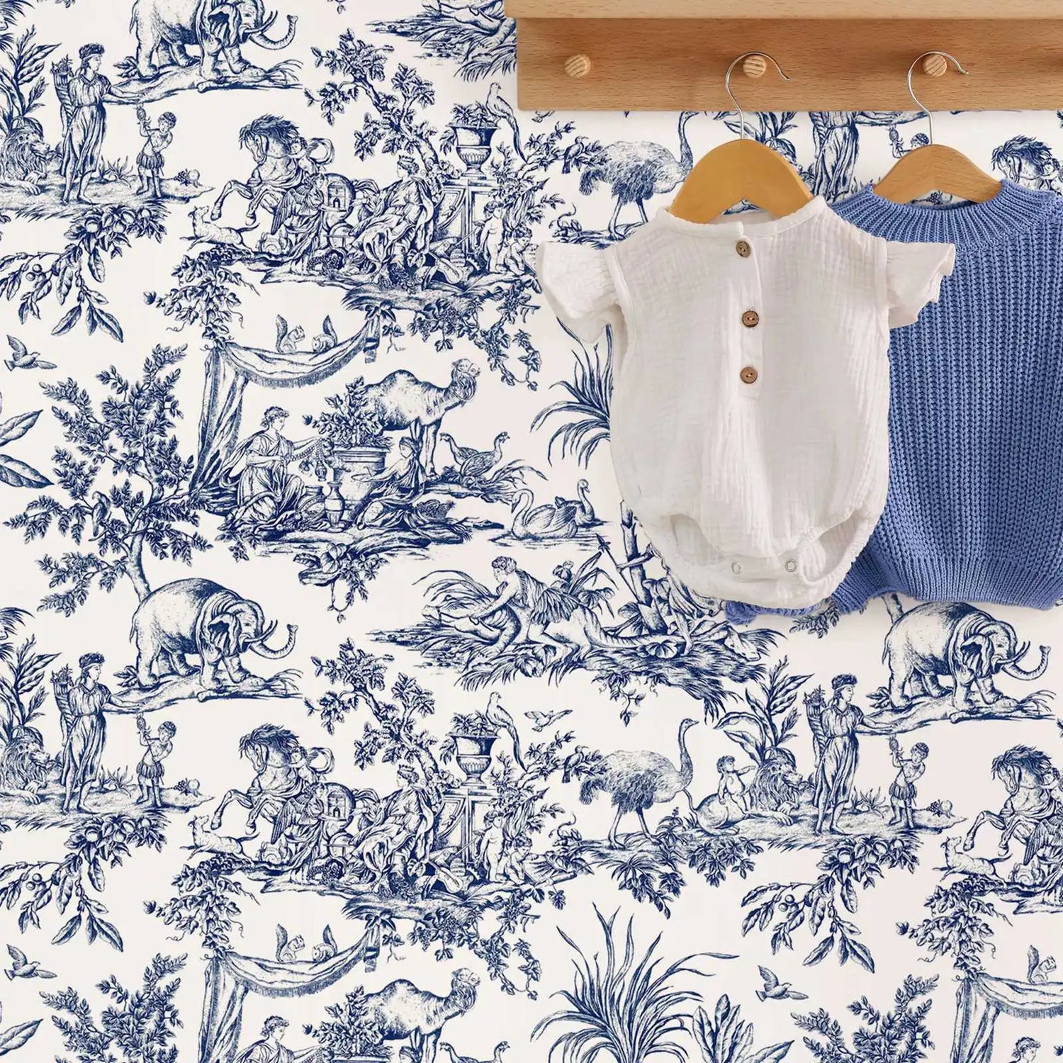 White baby romper and blue knitted cardigan on wooden hangers against a blue and white toile wallpaper background.