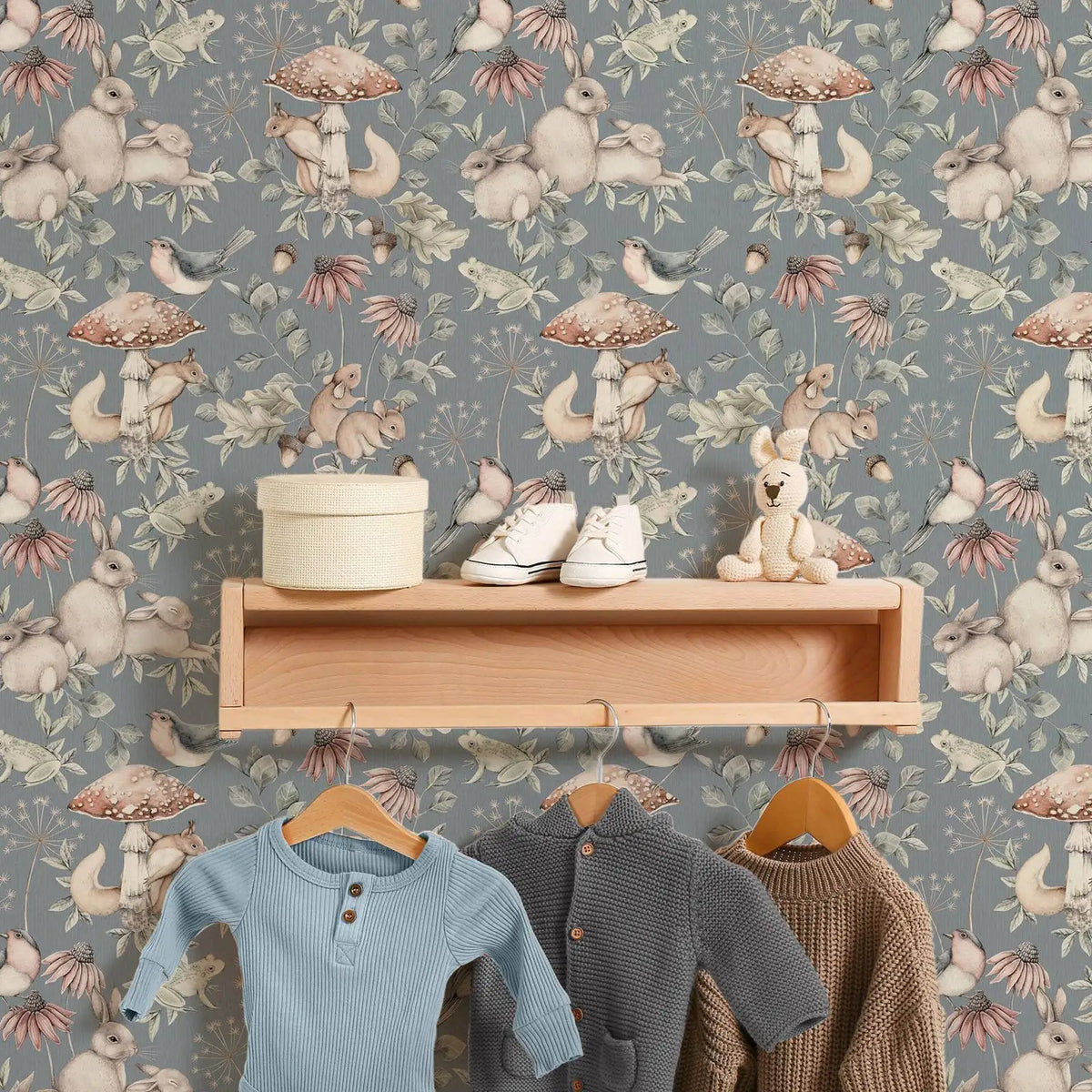 Children's clothing on hangers with a wooden shelf and toys against a patterned wallpaper.