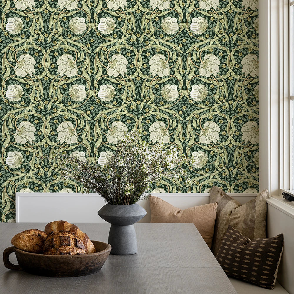 Room interior with floral-patterned wallpaper, table with bread and vase, and pillows on a bench.