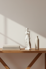 A minimalist interior scene with a white classical-style statue and a tall matte vase on a light wood table, alongside a stack of books, against a warm beige wall with soft diagonal sunlight and shadows.