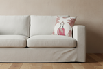 A square decorative throw pillow with a traditional Indian-style illustration of a woman in a red sari and gold crown, set against a warm beige background with pink palm leaves. The pillow is styled on a cream-colored fabric armchair next to a small wooden side table with yellow flowers, set against a textured brown wall.