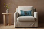 A cream-colored upholstered armchair with a light wood frame, styled with a blue patterned throw pillow, next to a small wooden side table holding a vase of yellow flowers against a textured brown wall.