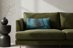 A cream-colored upholstered armchair with a light wood frame, styled with a blue patterned throw pillow, next to a small wooden side table holding a vase of yellow flowers against a textured brown wall.