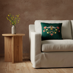 A light gray upholstered armchair with white turned legs sits in a styled room, featuring a dark green embroidered throw pillow with a bee and floral design. A wooden side table with flowers, a floor lamp, and paneled gray walls are visible.