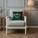 A light gray upholstered armchair with white turned legs sits in a styled room, featuring a dark green embroidered throw pillow with a bee and floral design. A wooden side table with flowers, a floor lamp, and paneled gray walls are visible.