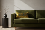 An olive green velvet sofa in a modern living room setting, styled with a yellow and blue embroidered throw pillow and a black side table with a vase of dried branches.