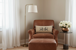 A brown leather armchair with a matching ottoman in a softly lit room, styled with a beige throw pillow featuring a black lion design. A brass floor lamp stands to the left, and a side table with white roses is on the right.