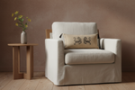 A cream-colored upholstered armchair with a matching side table holding a vase of yellow flowers, set against a textured brown wall on a light wood floor. The armchair features a beige throw pillow with a black embroidered pattern.