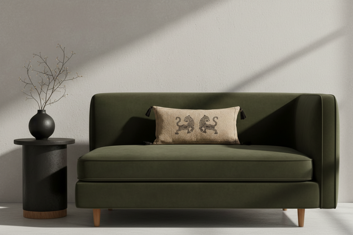 An olive green upholstered loveseat with tapered wooden legs, styled with a beige throw pillow featuring a black lion design, set against a textured light gray wall with dramatic diagonal shadows. A black side table with a black vase and dried branches stands to the left.
