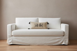 A white slipcovered loveseat sofa with a beige accent pillow featuring two dark gray lions, set against a neutral beige wall and light wood floor.