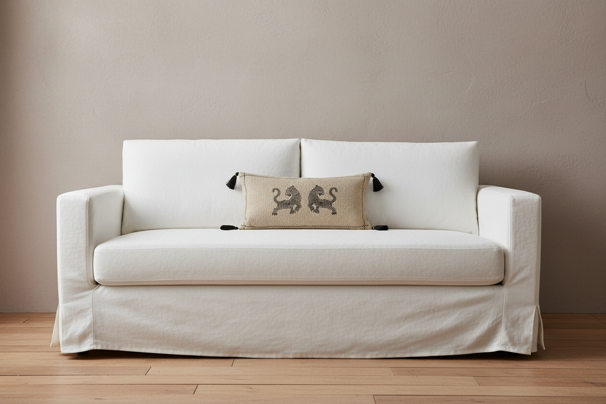 A white slipcovered loveseat sofa with a beige accent pillow featuring two dark gray lions, set against a neutral beige wall and light wood floor.