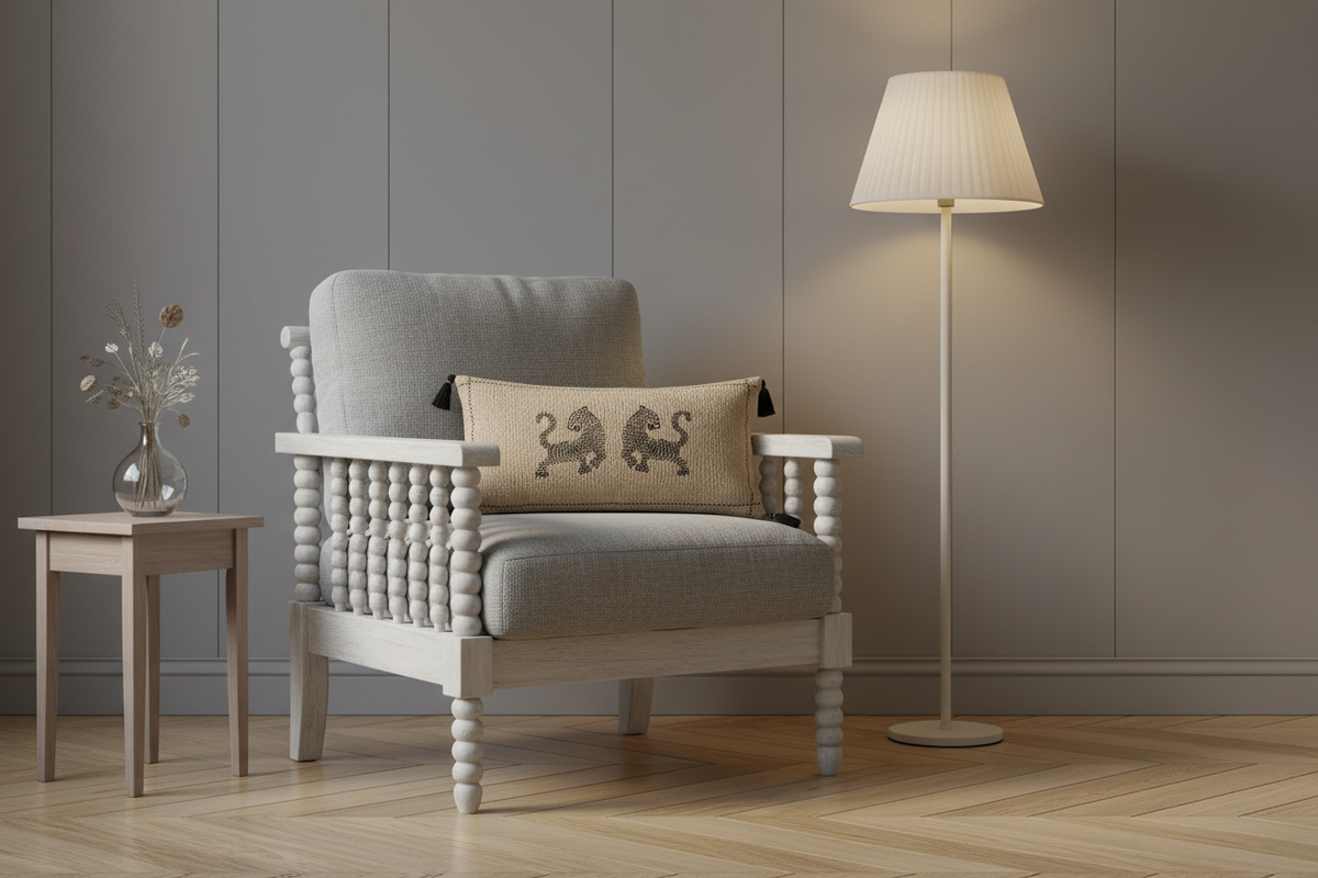 A light gray upholstered armchair with a white spindle-back frame, styled with a beige throw pillow featuring a black embroidered lion design. The chair is set in a modern room with a gray paneled wall, a wooden floor, a small side table with a vase, and a floor lamp.