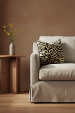 A beige slipcovered sofa in a styled room, featuring a black and beige zebra-patterned throw pillow, a wooden side table with a vase of yellow flowers, and a textured brown wall.