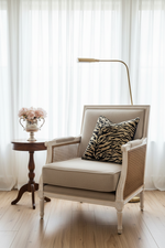 A beige upholstered armchair with woven cane sides and a black-and-beige zebra-pattern throw pillow, styled in a bright room with a wooden floor, sheer white curtains, a small side table, and a floor lamp.