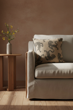 A beige upholstered armchair in a styled room setting, featuring a textured throw pillow with a dark animal print. A small wooden side table with a vase of yellow flowers stands to the left against a warm brown plaster wall and light wood flooring.