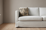 A light beige fabric slipcovered sofa with a textured throw pillow featuring a dark gray leopard print, set against a neutral beige wall and light wood flooring.