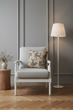 A light gray upholstered armchair with a decorative patterned throw pillow, set in a styled room with a gray paneled wall, a floor lamp, and a wooden side table.