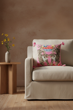 A pink decorative throw pillow with a tiger print and colorful floral details, styled on a white slipcovered sofa in a living room setting.