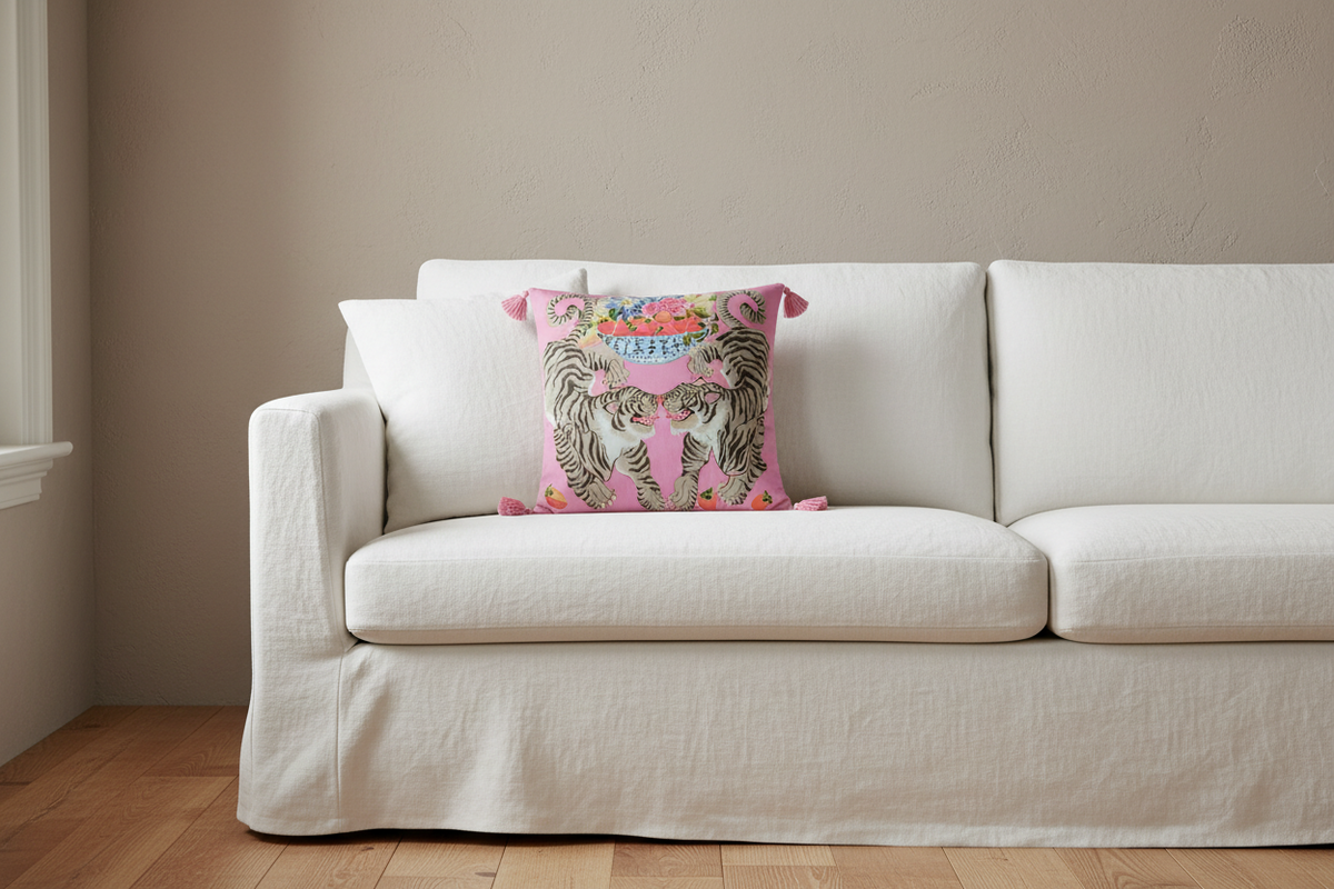 A pink decorative throw pillow with a tiger print and colorful floral details, styled on a white slipcovered sofa in a living room setting.