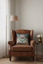 A brown leather wingback armchair in a styled room corner, featuring a colorful floral patterned throw pillow. A gold floor lamp stands to the left, and a small side table with white flowers is on the right.