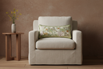 A cream-colored slipcovered loveseat sofa with a decorative floral accent pillow, set against a textured beige wall with a white vase of pampas grass on the left and a small wooden side table on the right.
