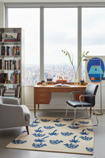Modern office with a desk, chair, turtle and palm tree rug, and bookshelf in front of large windows with a cityscape view.