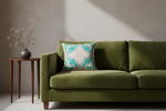 An olive green velvet sofa in a styled room setting, featuring a decorative teal and cream patterned throw pillow and a small wooden side table with dried flowers against a textured gray wall.