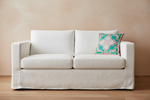 An olive green velvet sofa in a styled room setting, featuring a decorative teal and cream patterned throw pillow and a small wooden side table with dried flowers against a textured gray wall.