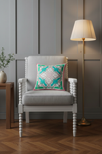 An olive green velvet sofa in a styled room setting, featuring a decorative teal and cream patterned throw pillow and a small wooden side table with dried flowers against a textured gray wall.