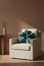 A cream-colored upholstered armchair in a warm, sunlit room, styled with a large teal succulent-shaped throw pillow. A small wooden side table with a vase of yellow flowers sits to the left against a textured brown wall and light wood flooring.