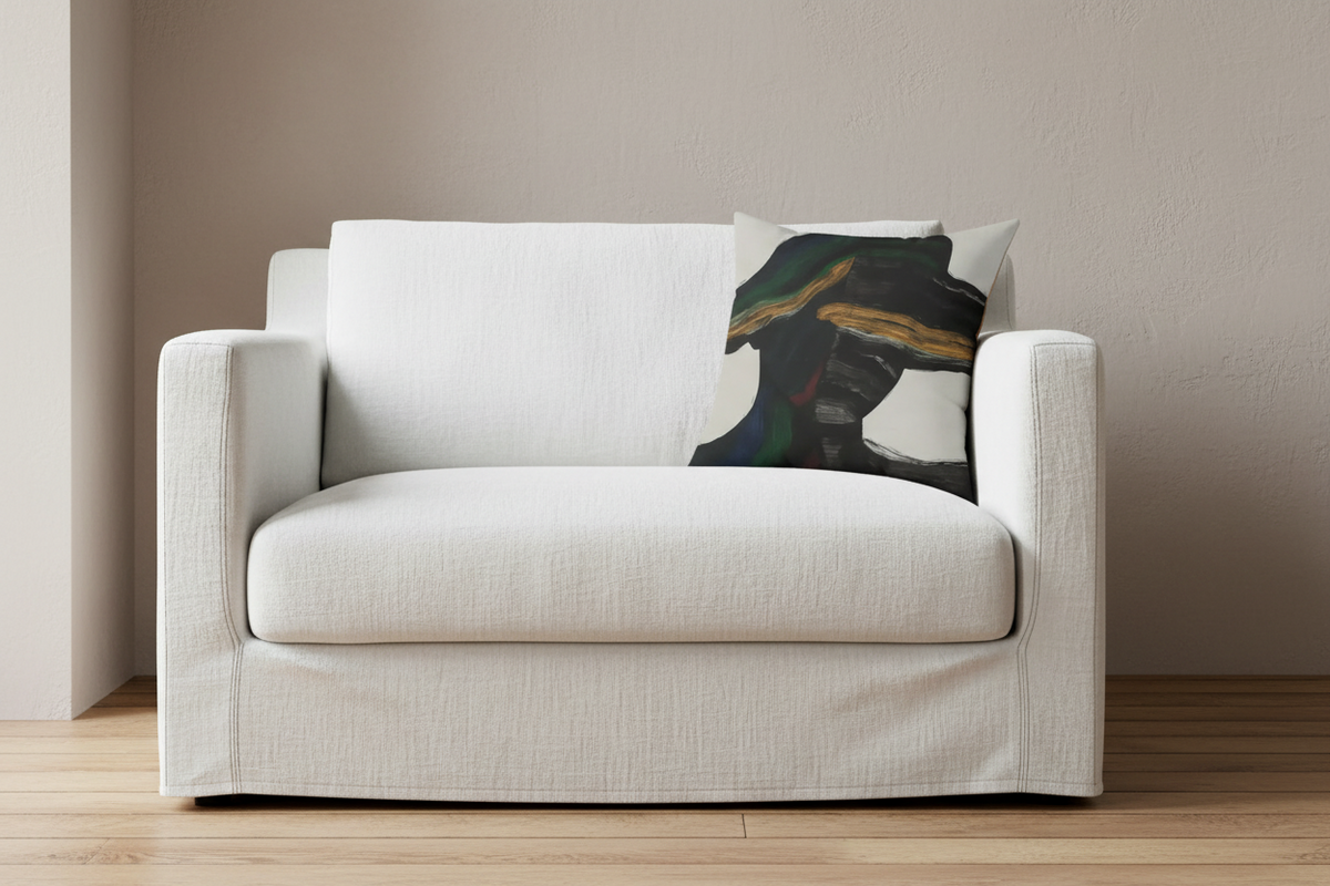 A light beige upholstered armchair with a matching slipcover, shown in a minimalist room setting with a textured beige wall and light wood flooring. A decorative throw pillow with an abstract black, green, and gold design rests on the right side of the chair.