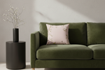 An olive green velvet sofa in a modern living room setting, styled with a light pink floral throw pillow and a black side table holding a black vase with dried branches.